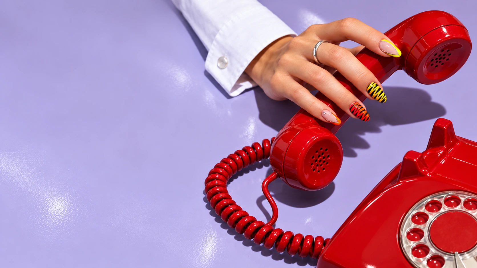 Campagna di concept: una mano con nail art fluo solleva un telefono rosso su un piano lilla.